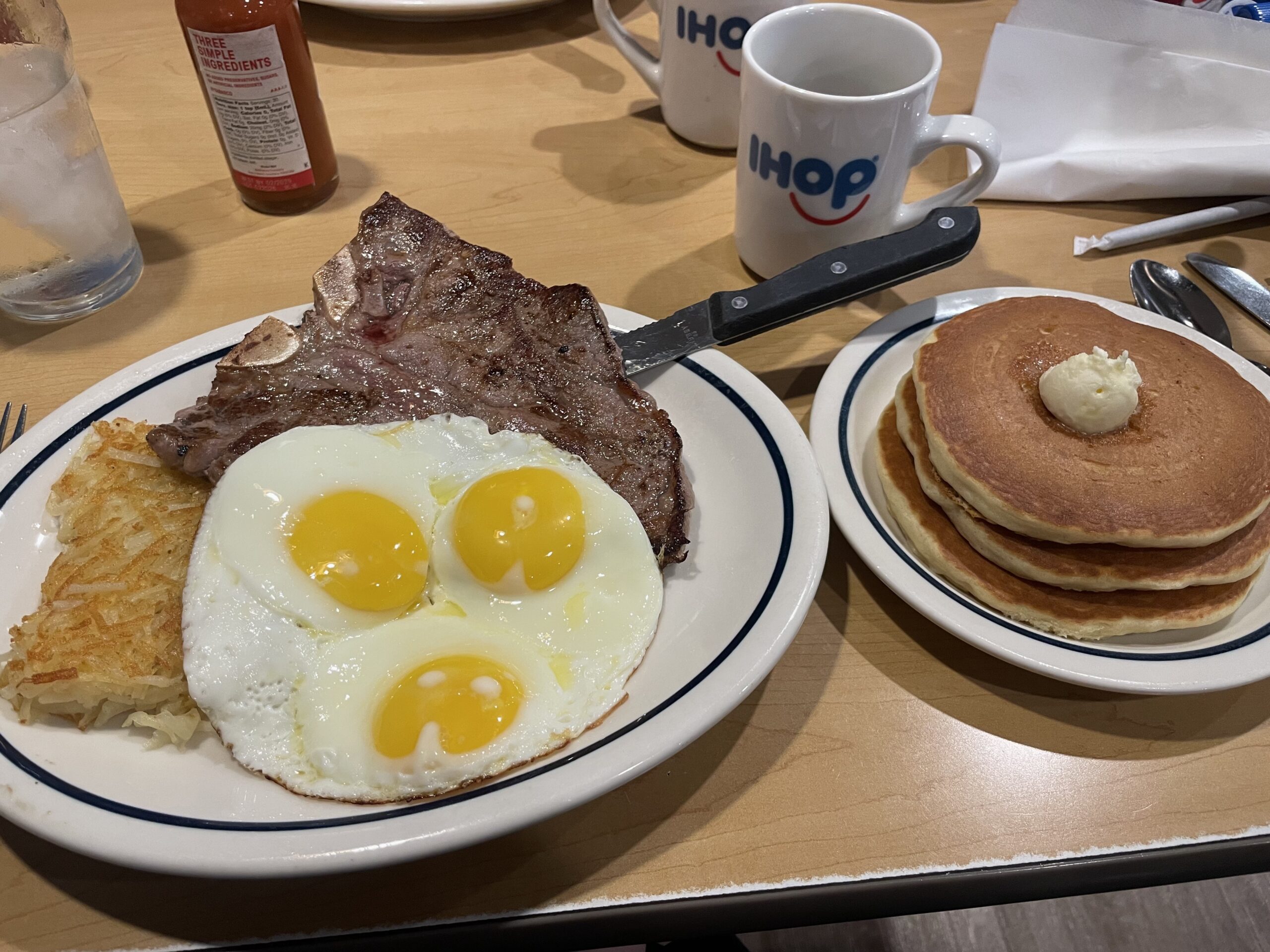 Large plate with hash browns, steak, and three eggs beside a smaller plate with a stack of three pancakes, beside a mug with the IHOP logo.