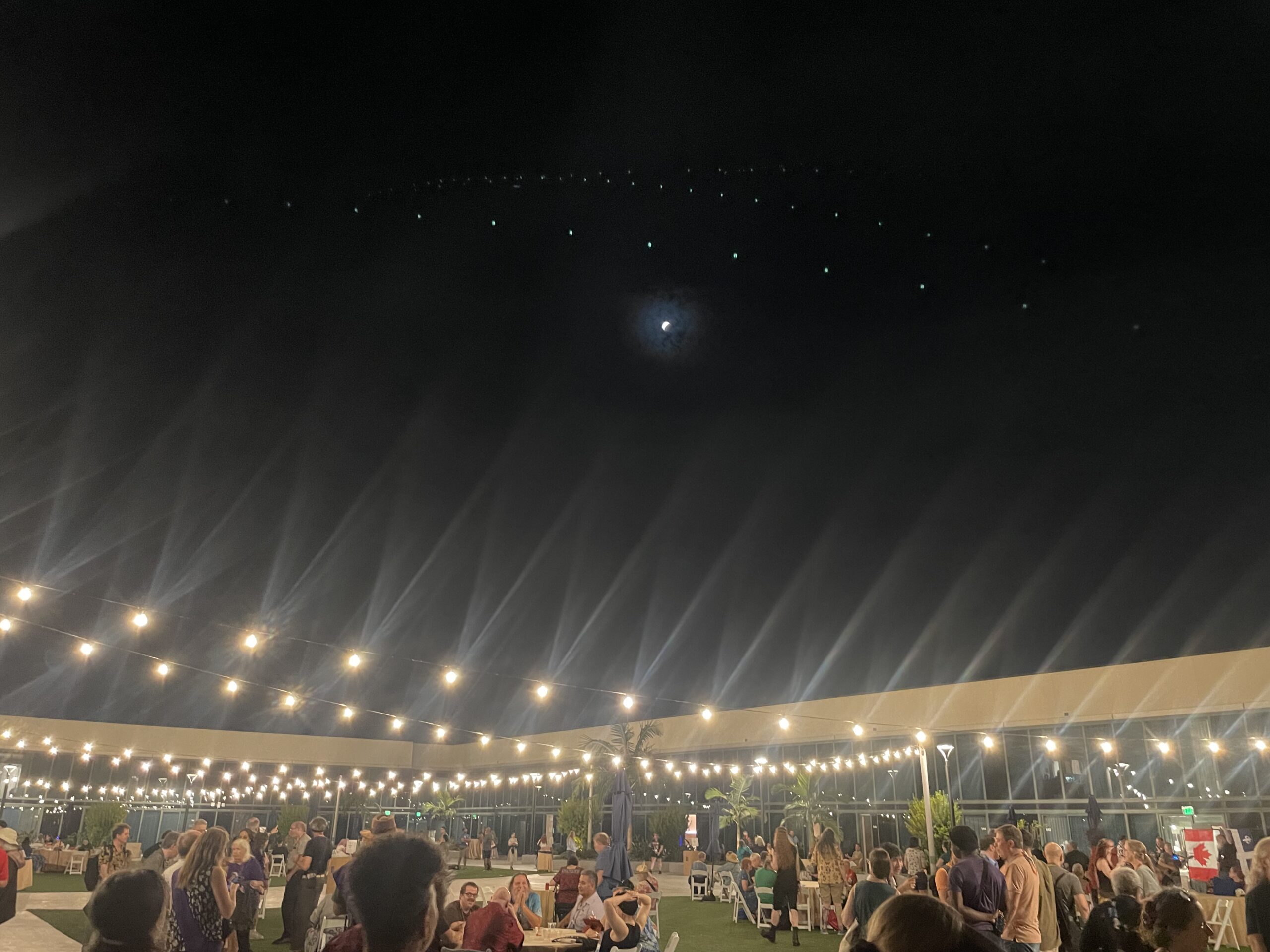 An outdoor patio with lots of people under fairy lights and a tiny moon in partial eclipse above